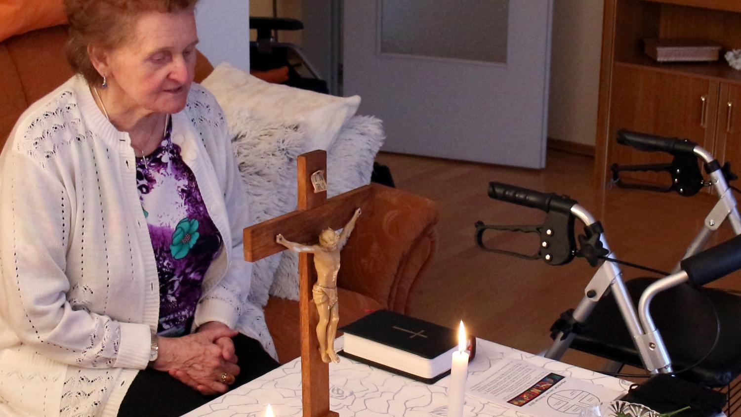 Eine ältere Person sitzt in einem Wohnzimmer an einem Tisch, der mit einer weißen, gemusterten Tischdecke bedeckt ist. Auf dem Tisch stehen ein hölzernes Kruzifix, zwei brennende Kerzen, eine Bibel und ein kleiner Zweig in einer Glasschale. Neben dem Tisch befindet sich ein Rollator. In der Umgebung sind ein Sofa mit Kissen und ein Regal zu sehen.
