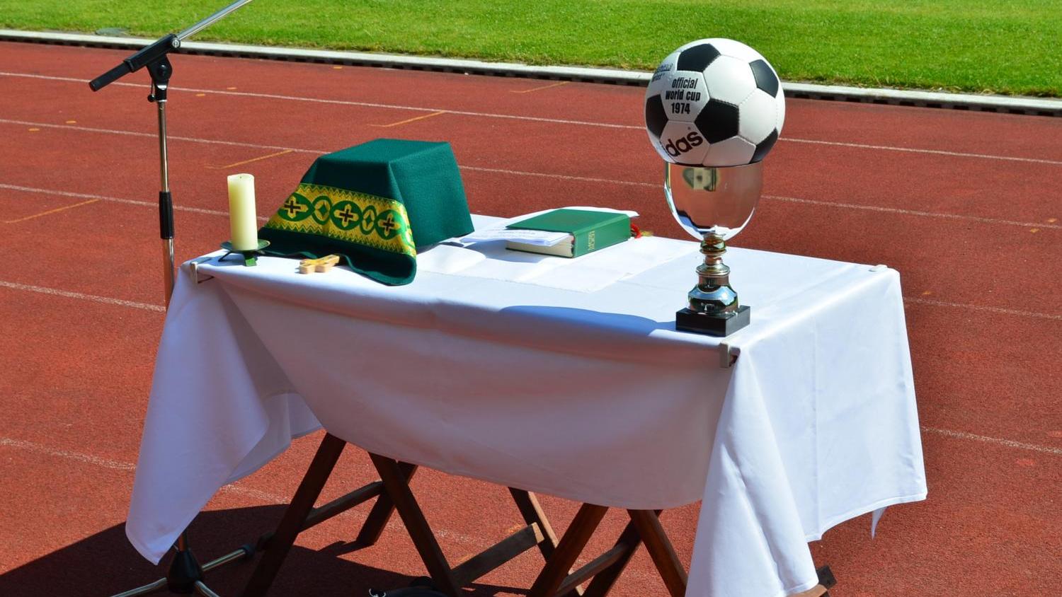 Vor dem grünen Rasen eines Stadions ist auf der roten Laufbahn ein provisorischer Altar aufgebaut. Auf dem weiß gedeckten Tisch liegen ein grünes liturgisches Tuch, eine Kerze, ein kleines Holzkreuz und ein liturgisches Buch. Links steht ein Mikrofonständer für eine Freiluftandacht oder einen Gottesdienst. Rechts auf dem Tisch befindet sich ein silberner Pokal, auf dem ein Fußball mit der Aufschrift „Official World Cup 1974“ platziert ist. Die Szene verbindet christliche Symbole mit dem Umfeld eines Sportplatzes.